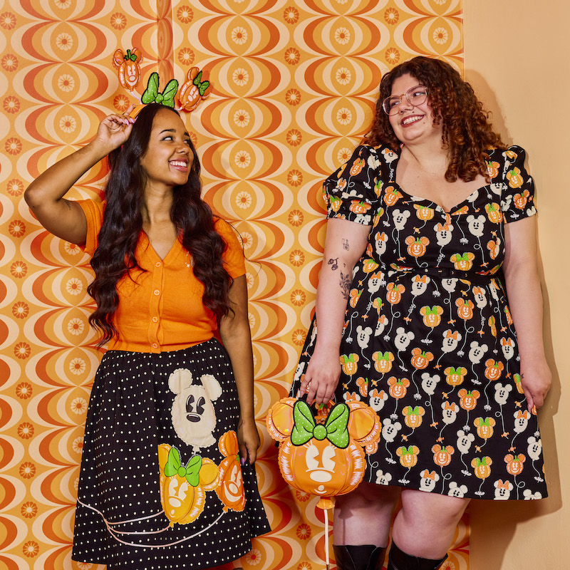 Two women laughing together, one wearing the Stitch Shoppe Minnie Mouse Pumpkin Balloon Domonique Cardigan and the Minnie Mouse Pumpkin Balloon Sandy Skirt, and the other wearing the Stitch Shoppe Minnie Mouse Pumpkin Balloon Allison Dress against a psychedelic orange background 